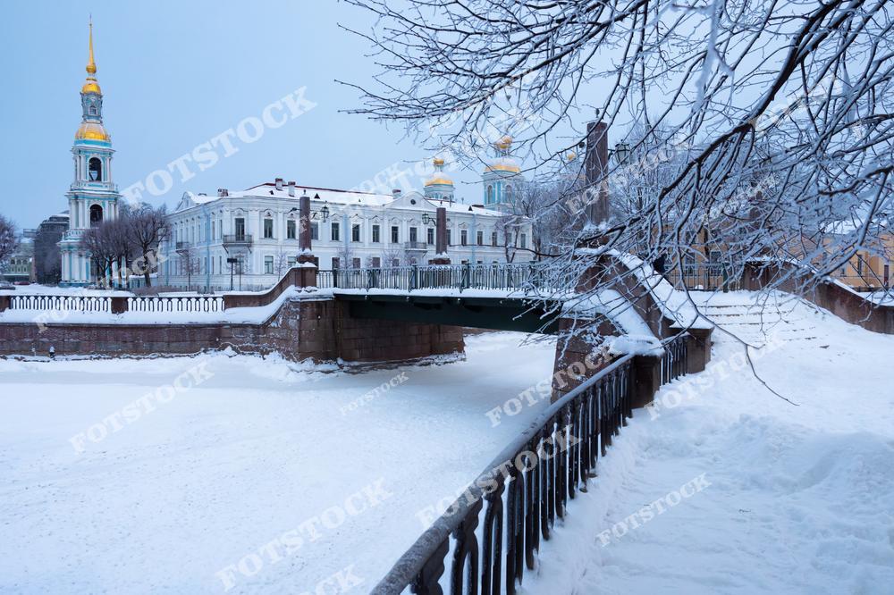 На снимке изображён Николо-Богоявленский морской собор — один из красивейших храмов Санкт-Петербурга. Вид открывается с набережной канала, вдоль которого тянутся ограды и покрытые снегом деревья. Хорошо видна колокольня собора с золотым шпилем, а также мост, перекинутый через канал. Это историческая часть города, известная своей архитектурой и атмосферой. Зимний пейзаж подчёркивает спокойствие и величие православной святыни.